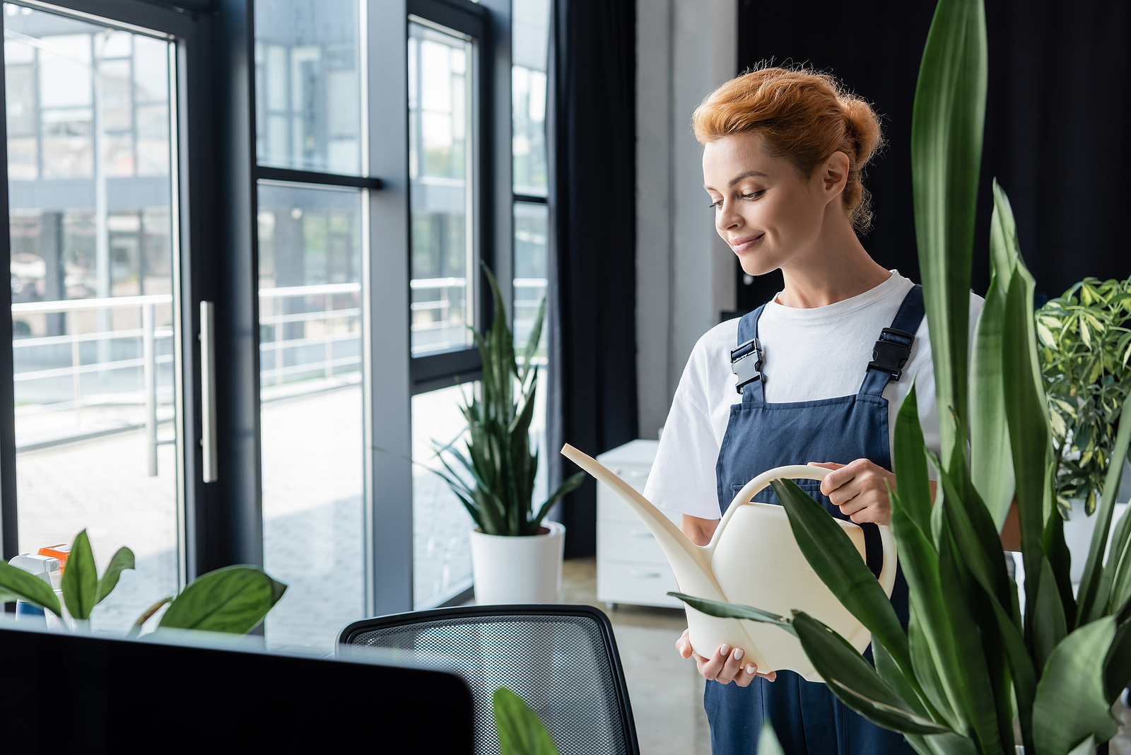 Eine junge Frau gießt mit Hilfe einer Gießkanne die Pflanzen im Büro die sich in der Nähe der Fenster befinden