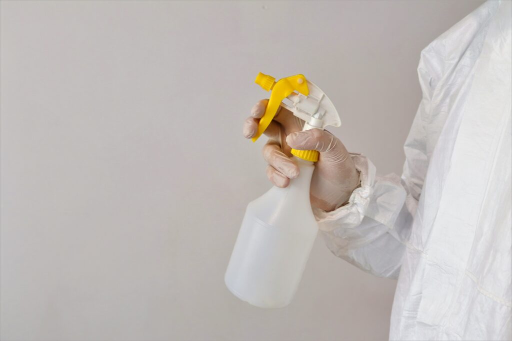 Symbolbild für Schimmel überstreichen: Eine Person in einem Schutzanzug hält eine Sprühflasche vor einer weißen Wand in der rechten Hand