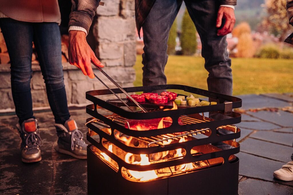 im Höfats Cube Feuerkorb in schwarz brennt ein feuer. Über dem Feuer ist ein Grillrost auf dem Fleisch und Gemüse grills. Hinter dem Grill stehen personen eine Person wendet das Fleisch was auf dem Grillrost liegt