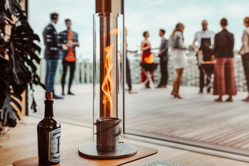 Die SPIN 90 Tischfeuer grau von Höfats  steht auf einem Tisch auf einer Dachterasse. Im Tzylindrischen Tischfeuer brennt eine Flamme. Im Hintergrund stehen Menschen um Höfats Grills und neben dem Tischfeuer steht eine Flasche Ferdinans Gin.