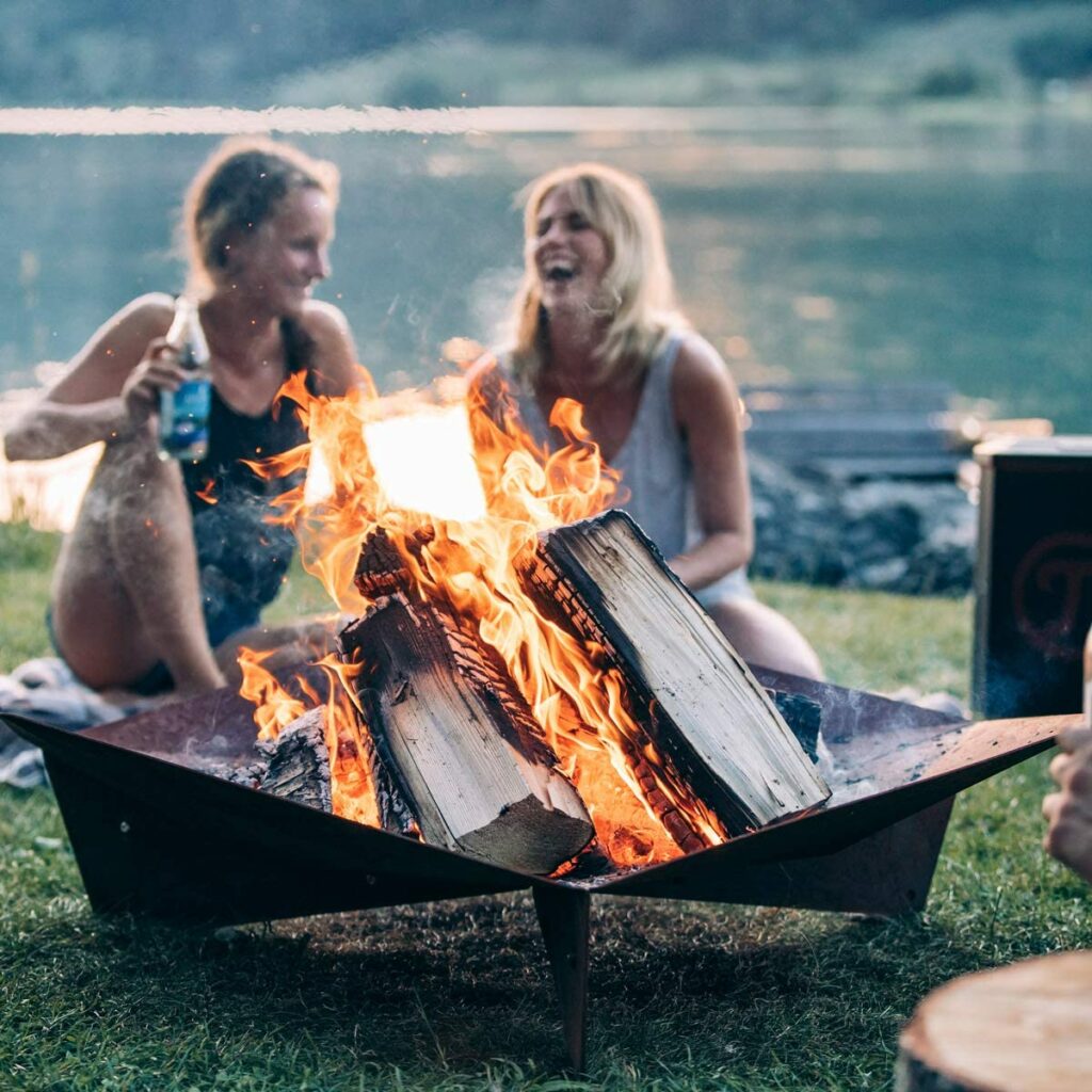 Die Höfts triple Feurschale mit brennenden Holzscheiten an einem See. Hinter der feuerschale sitzen zwei Frauen die lachen