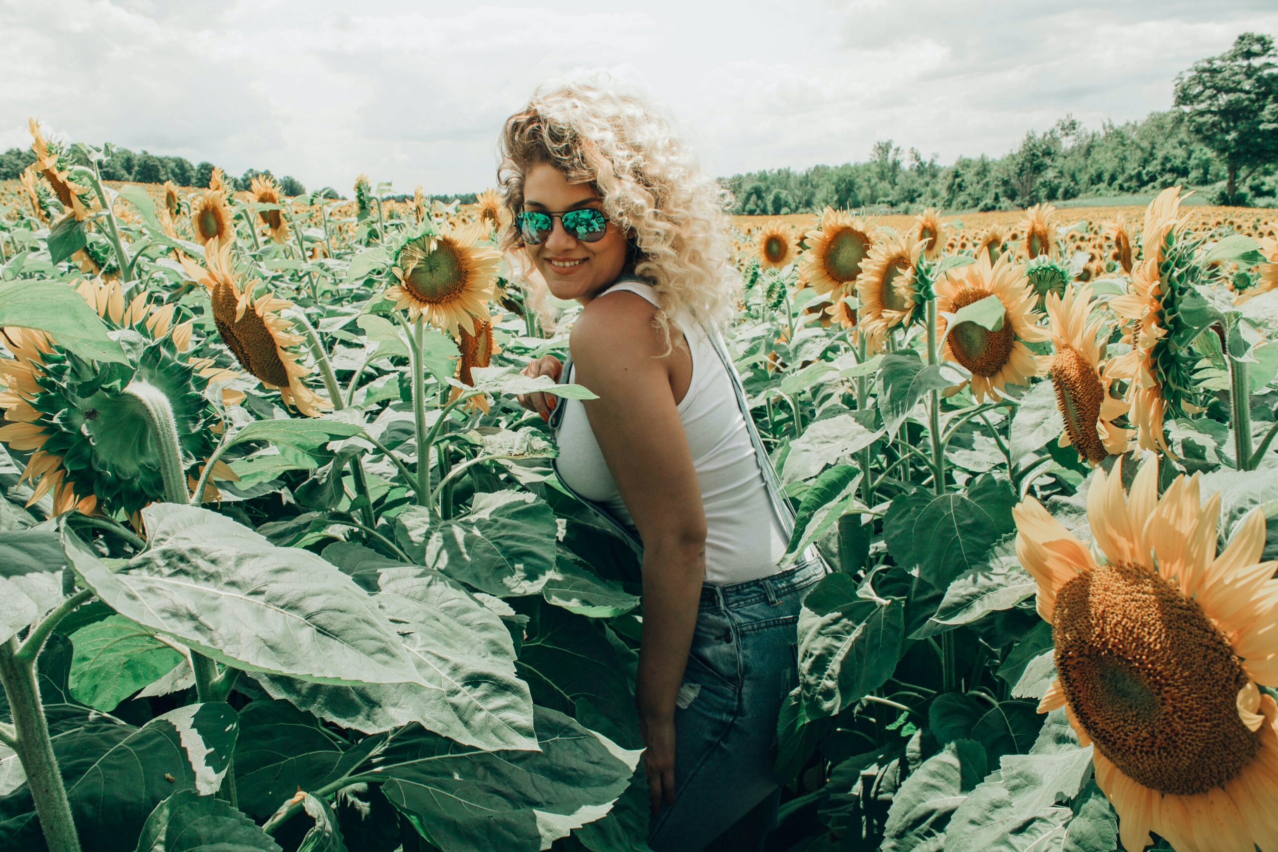 Eine junge Frau mit blonden Haaren und Sonnnenbrille läuft lächelt durch ein Beet mit Sonnenblumen