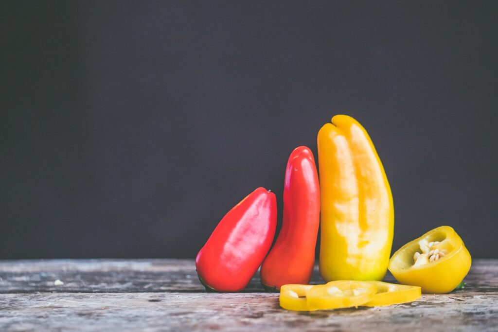 3 Chili-Schoten, zwei rote eine gelbe auf einem Tisch vor einer grauen wand. Chili-Anbau sind die perfekte Wahl für alle, die einen Schuss Geschmack und Farbe in ihren Garten oder auf ihre Terrasse bringen möchten
