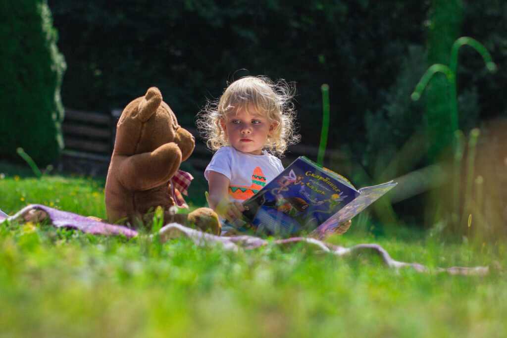 Ein kleines Kind sind auf einer Decke im Garten und ließt aus einem Kinderbuch der Teddybär der neben dem Kind sitz vor.