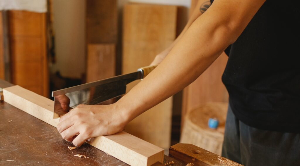 Eine Person sägt mit einer Handsäge durch ein Stück Holz das auf einer Werkbank liegt.
