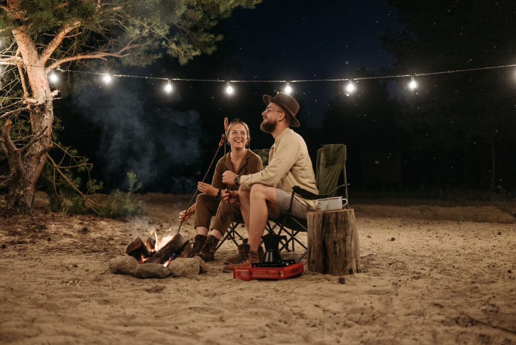 Ein junges Paar sitzt bei Nacht an einer Feuerstelle am Strand und grillt mit Spießen über einem Lagerfeuer