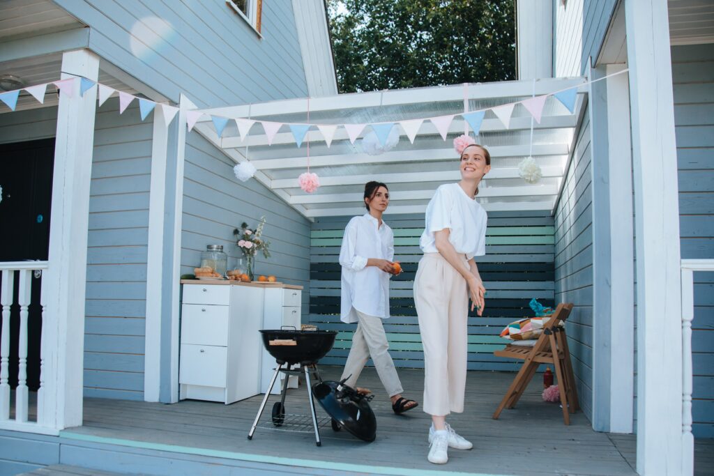 Zwei junge Frauen stehen unter dem geschmücktem Vordach ihres Hauses und bereiten eine Gartenparty vor.