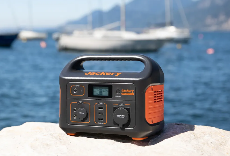 Jackery Explorer Powerstation steht auf einem Stein am Ufer eines Meeres. Im verschwommenen Hintergrund stehen Segelschiffe im Wasser