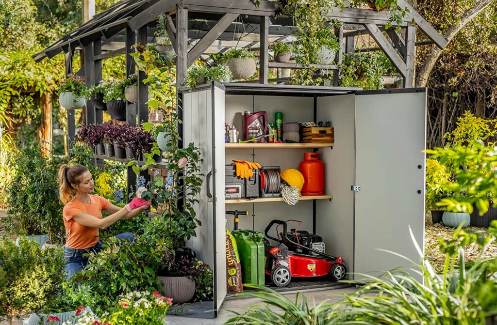 Die Keterbox Premier Tall Gartenlaube gartenhaus steht geöffnet in einem Garten gefüllt mit unterschiedlichen Gartengeräten